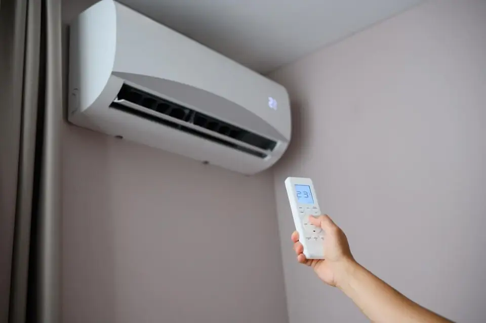 Biała jednostka wewnętrzna klimatyzacji na ścianie, w dole kadru dłoń z pilotem pokazującym 23 stopnie.