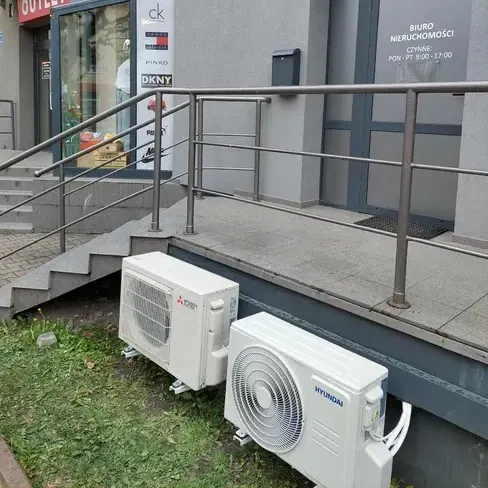 Dwie jednostki zewnętrzne klimatyzacji, Mitsubishi Electric i Hyundai, ustawione na stopach montażowych przy wejściu do lokalu usługowego.