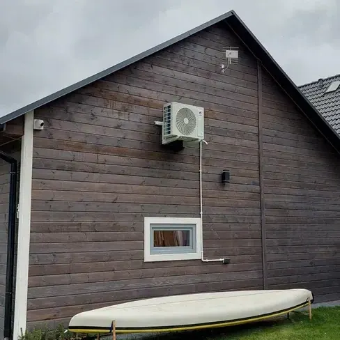 Jednostka zewnętrzna klimatyzacji zamontowana na wspornikach na ciemnej drewnianej elewacji szczytowej budynku, z prowadzonym pionowo białym korytem instalacyjnym.