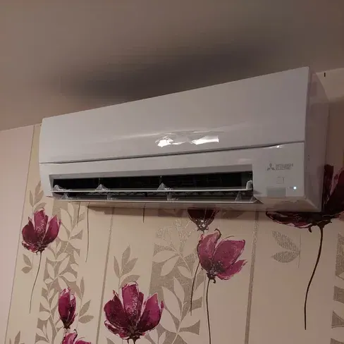 Biała jednostka wewnętrzna klimatyzacji Mitsubishi Electric zamontowana wysoko na ścianie z tapetą w duże bordowe kwiaty.